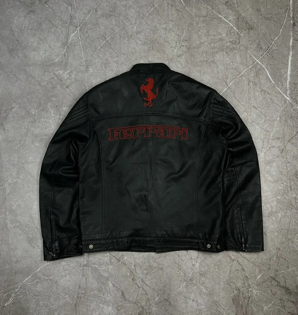 Back view of the black leather Ferrari jacket showing the red embroidered prancing horse logo and FERRARI text on a marble background.