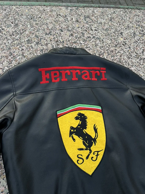 Back view of the black Ferrari F1 leather jacket showing the large red "Ferrari" text and the yellow Scuderia Ferrari shield.