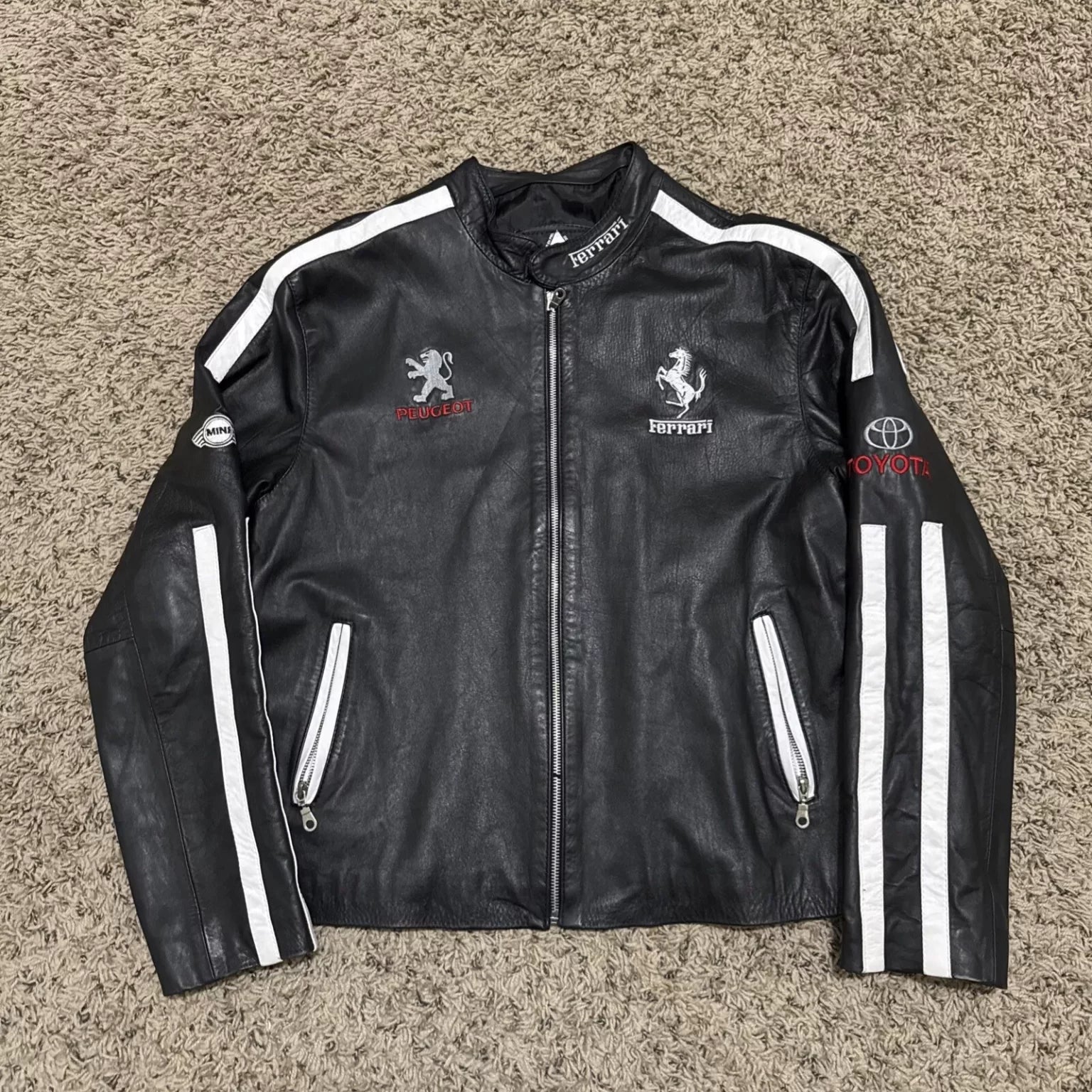 Full front view of a black leather multi-brand motorsport jacket with white stripes, featuring Peugeot, Ferrari, Toyota, and MINI logos.